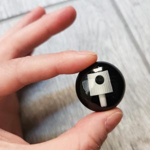 May include: A small, circular black button featuring a white spray paint nozzle graphic centered on its surface. The design is glossy and smooth, showcasing a detailed illustration of a standard aerosol cap used for graffiti and street art applications.