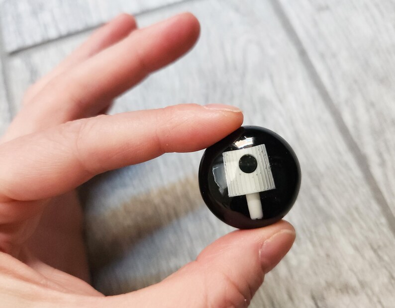 May include: A small, circular black button featuring a white spray paint nozzle graphic centered on its surface. The design is glossy and smooth, showcasing a detailed illustration of a standard aerosol cap used for graffiti and street art applications.