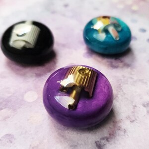 May include: Three circular buttons with a smooth, glossy finish in black, teal, and purple. Each button features a metallic spray paint nozzle cap embedded in the center, with the purple button showcasing a gold-toned nozzle and the others silver.