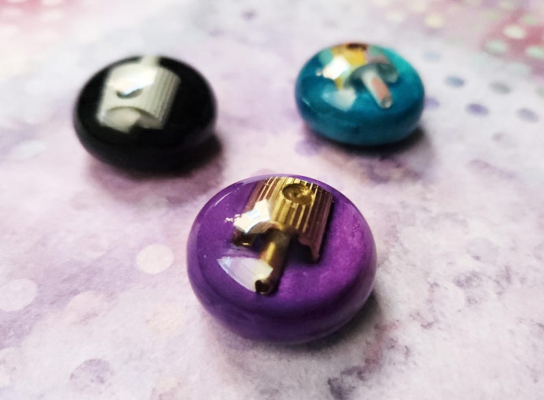 May include: Three circular buttons with a smooth, glossy finish in black, teal, and purple. Each button features a metallic spray paint nozzle cap embedded in the center, with the purple button showcasing a gold-toned nozzle and the others silver.