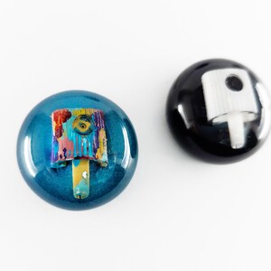 May include: Two circular spray paint caps. One cap is metallic blue with a multi-colored paint-splattered nozzle. The other cap is solid black with a plain white nozzle. Both caps feature a smooth, glossy finish and a rounded, dome-like shape.