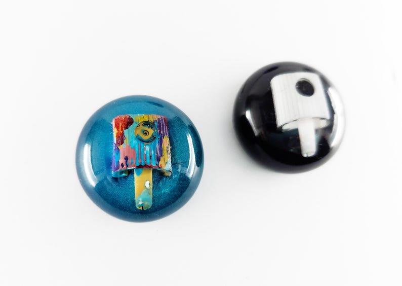 May include: Two circular spray paint caps. One cap is metallic blue with a multi-colored paint-splattered nozzle. The other cap is solid black with a plain white nozzle. Both caps feature a smooth, glossy finish and a rounded, dome-like shape.