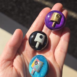 May include: Three circular resin pins featuring embedded spray paint caps. The top pin is metallic purple with a gold cap, the middle is solid black with a white cap, and the bottom is glittery blue with a multi-colored patterned spray cap.