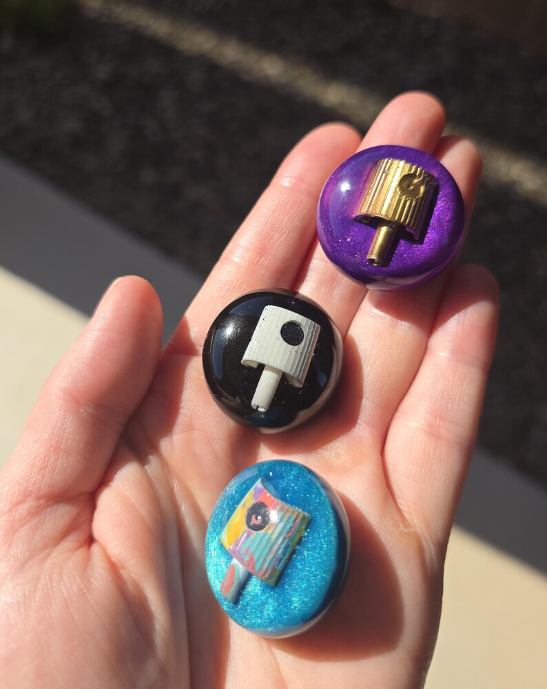 May include: Three circular resin pins featuring embedded spray paint caps. The top pin is metallic purple with a gold cap, the middle is solid black with a white cap, and the bottom is glittery blue with a multi-colored patterned spray cap.