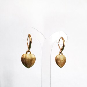 May include: A pair of gold heart-shaped earrings with a textured surface. The earrings are hanging from a clear plastic display stand.