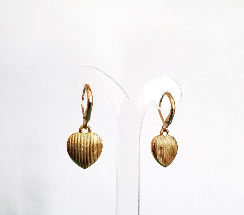 May include: A pair of gold heart-shaped earrings with a textured surface. The earrings are hanging from a clear plastic display stand.