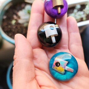 May include: Three circular pins featuring embedded spray paint nozzles. One pin is purple with a gold nozzle, one is black with a white nozzle, and one is teal with a multi-colored nozzle. Each pin has a smooth, glossy, rounded surface finish.