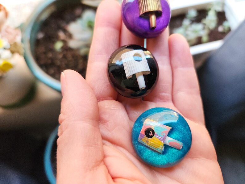 May include: Three circular pins featuring embedded spray paint nozzles. One pin is purple with a gold nozzle, one is black with a white nozzle, and one is teal with a multi-colored nozzle. Each pin has a smooth, glossy, rounded surface finish.