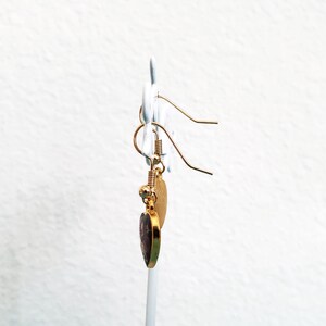 May include: Gold heart-shaped earrings with a purple stone in the center. The earrings are hanging from a gold hook.