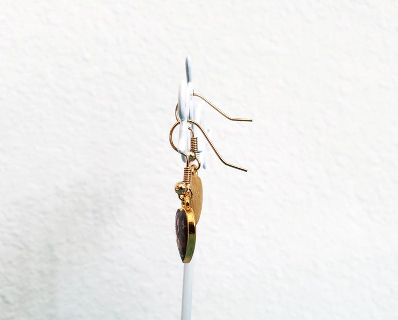 May include: Gold heart-shaped earrings with a purple stone in the center. The earrings are hanging from a gold hook.