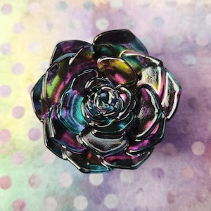 May include: A metallic rose-shaped object featuring an iridescent finish with shifting hues of deep purple, blue, and green. The petals are layered in a circular pattern, creating a reflective surface that catches light across its curved edges.