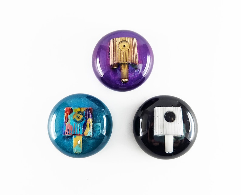 May include: Three circular resin objects featuring embedded spray paint caps. One is purple with a gold-toned cap, one is teal with a multi-colored paint-splattered cap, and one is black with a white cap. Each piece has a smooth, glossy finish.