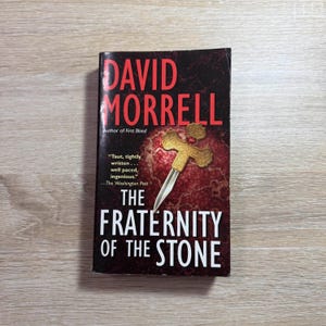 May include: A hardcover book titled "The Fraternity of the Stone" by David Morrell. The cover is dark red with gold accents, featuring a cross and dagger design. The book is on a light wood surface.