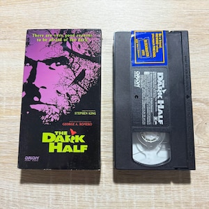May include: A VHS tape and its case for the movie "The Dark Half" by Stephen King. The case features a purple and black design with the movie title in yellow and green. The VHS tape is black with a Blockbuster Video sticker.