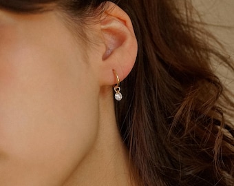 May include: A close-up of a person's ear with a small gold hoop earring with a single clear stone dangling from it.