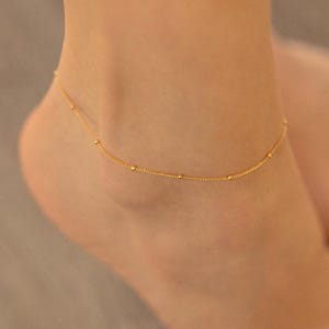 Puede incluir: Un delicado tobillo de oro con pequeñas cuentas redondas. El tobillo está en el tobillo de una persona.