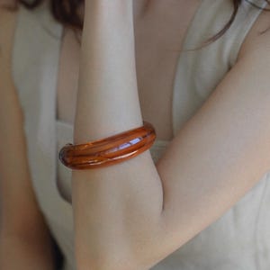 Könnte beinhalten: Ein durchscheinendes, bernsteinfarbenes Armreif-Armband. Das Armband ist ein dickes, abgerundetes Band mit glänzendem Finish. Die warmen Brauntöne des Armbands kontrastieren mit dem Hautton der Person, die es trägt. Das Armband ist ein auffälliges Schmuckstück.