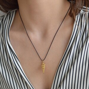 May include: A gold-toned pendant shaped like a spiral seashell suspended from a thin black cord necklace. The pendant features a textured, conical design with a pointed base, hanging centrally against a white and black vertical striped top.