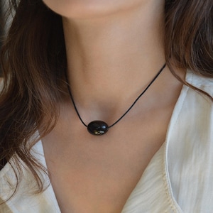 Peut inclure: Un pendentif noir de forme ovale suspendu à un fin cordon noir. Le pendentif présente une surface lisse, polie et réfléchissante. Le collier repose autour du cou, arborant un design simple avec un point focal unique au centre.