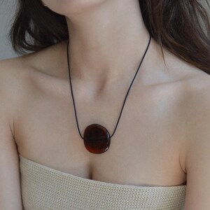 Op de afbeelding: Een ketting met een donker amberkleurige hanger. De hanger heeft een gladde, afgeronde vorm en is bevestigd aan een dun zwart koord. De ketting wordt gedragen over een top in neutrale tinten. De sieraden zijn eenvoudig en elegant.