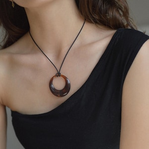 Op de afbeelding: Een ketting met een donkerbruine, cirkelvormige hanger en een zwart koord. De hanger heeft een schildpadpatroon en wordt gedragen op een zwarte top. Een eenvoudig, elegant accessoire.