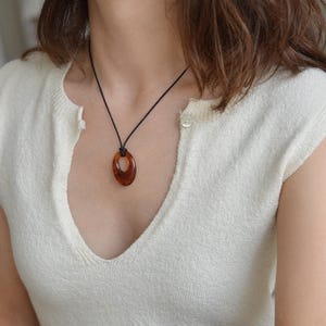 May include: A brown oval pendant necklace with a black cord. The pendant is a warm, translucent brown color. The necklace is worn over a cream-colored, short-sleeved top with a V-neck.