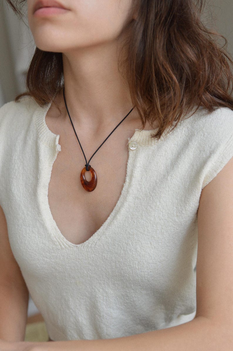 May include: A brown oval pendant necklace with a black cord. The pendant is a warm, translucent brown color. The necklace is worn over a cream-colored, short-sleeved top with a V-neck.