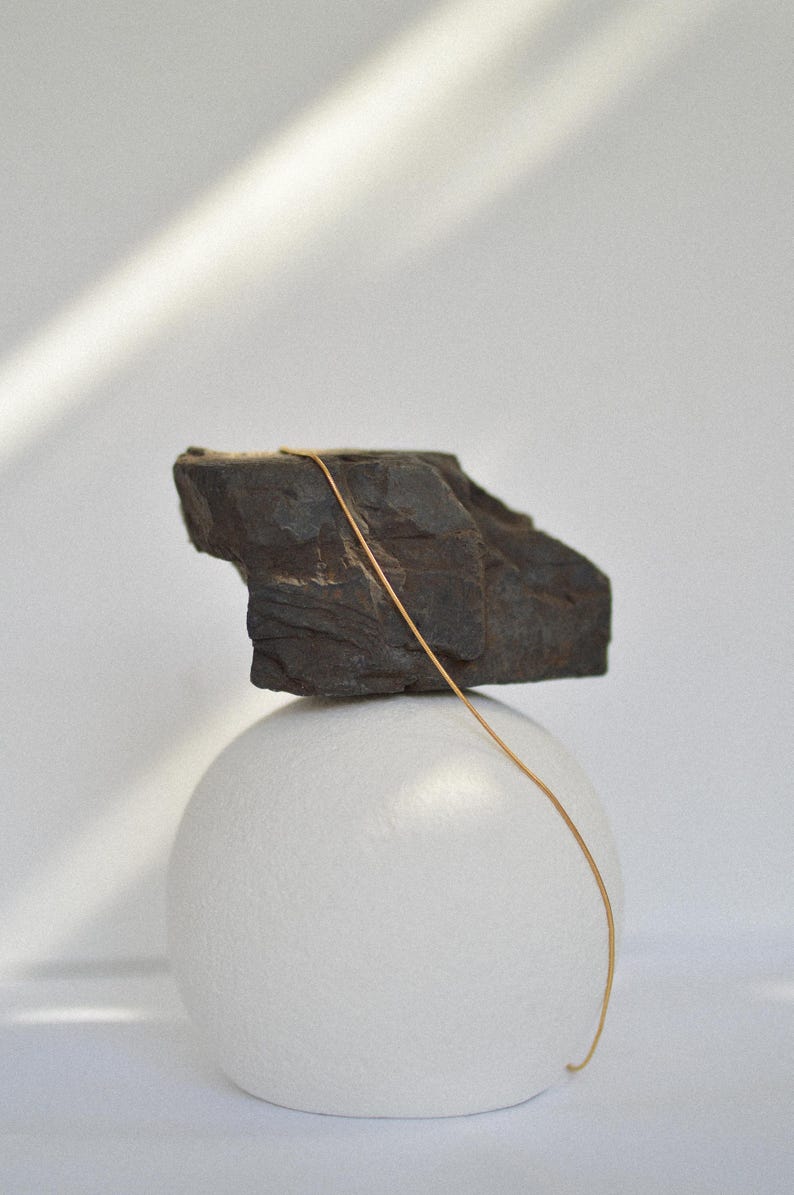 May include: A gold chain necklace draped over a dark grey rock, which is balanced on top of a white sphere.