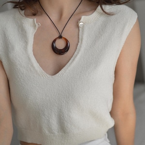 Op de afbeelding: Een cr&egrave;mekleurige mouwloze trui met een diepe V-hals. Een donkerbruine, cirkelvormige hangerketting hangt aan een zwart koord. De ketting heeft een schildpadpatroon. De trui heeft twee kleine knopen bij de halslijn.