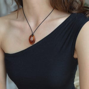May include: A brown oval pendant necklace with a black cord. The pendant is a translucent amber color. The necklace is worn over a black one-shoulder top.