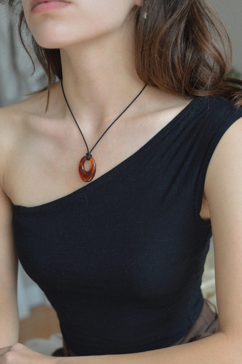 May include: A brown oval pendant necklace with a black cord. The pendant is a translucent amber color. The necklace is worn over a black one-shoulder top.