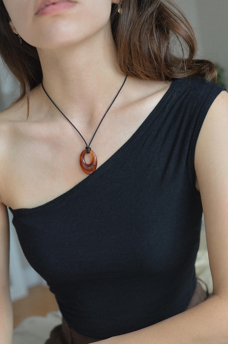 May include: A brown oval pendant necklace with a black cord. The pendant has a layered design with a glossy finish. The necklace is worn against a black one-shoulder top.