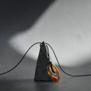 May include: A pendant necklace featuring an oval-shaped, amber-colored glass pendant. The pendant is suspended from a black cord and rests on a dark gray, triangular stone. The background is a gradient of gray and black.