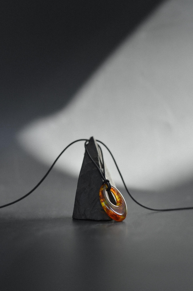 May include: A pendant necklace featuring an oval-shaped, amber-colored glass pendant. The pendant is suspended from a black cord and rests on a dark gray, triangular stone. The background is a gradient of gray and black.