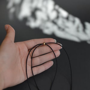 May include: A black necklace with a gold-colored bead. The necklace is held in a hand against a dark background. The necklace appears to be made of a thin, cord-like material.