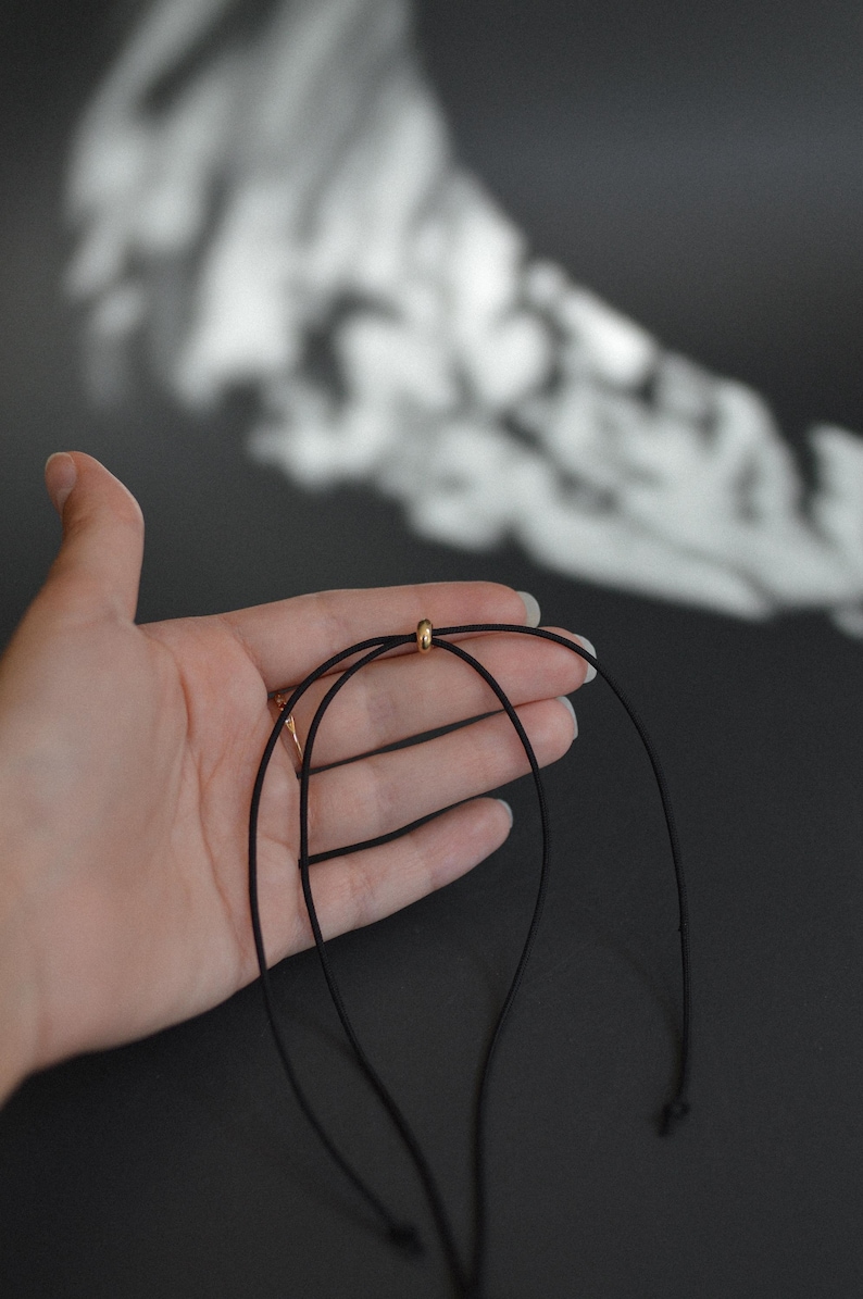 May include: A black necklace with a gold-colored bead. The necklace is held in a hand against a dark background. The necklace appears to be made of a thin, cord-like material.