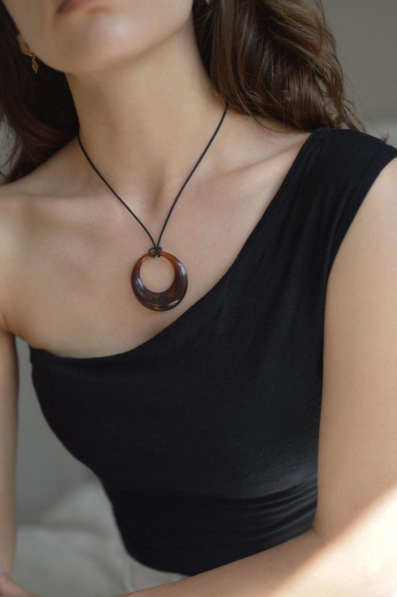 Op de afbeelding: Een ketting met een donkerbruine, cirkelvormige hanger en een zwart koord. De hanger heeft een schildpadpatroon en wordt gedragen op een zwarte top. Een eenvoudig, elegant accessoire.