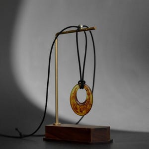 May include: A pendant necklace with a circular, amber-colored resin pendant. The pendant hangs from a black cord and is displayed on a gold-colored stand with a wooden base. The background is a neutral gray.