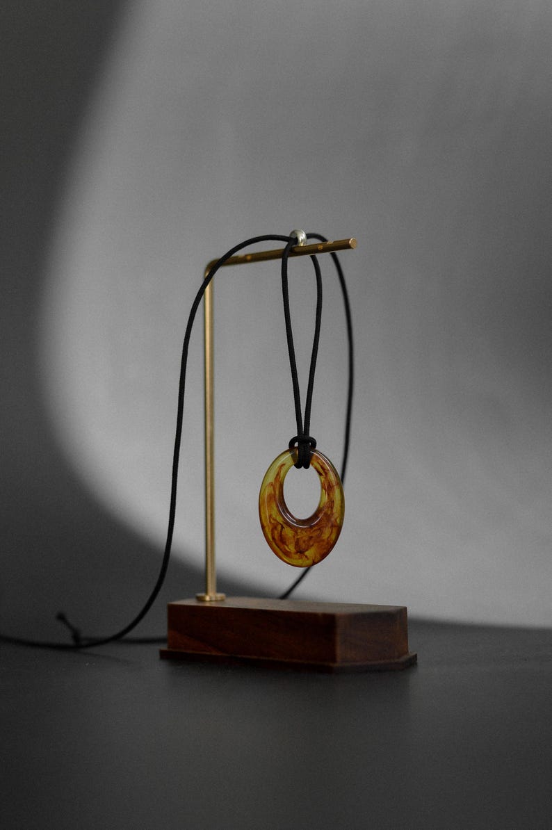May include: A pendant necklace with a circular, amber-colored resin pendant. The pendant hangs from a black cord and is displayed on a gold-colored stand with a wooden base. The background is a neutral gray.