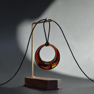 Op de afbeelding: Een hangerketting met een grote, cirkelvormige amberkleurige hars hanger die aan een zwart koord hangt. De hanger wordt getoond op een goudkleurige standaard met een houten voet, tegen een donkere achtergrond.