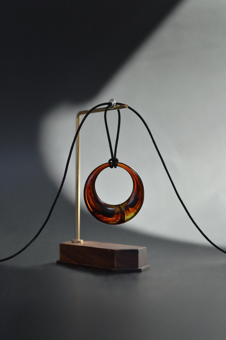 Op de afbeelding: Een hangerketting met een grote, cirkelvormige amberkleurige hars hanger die aan een zwart koord hangt. De hanger wordt getoond op een goudkleurige standaard met een houten voet, tegen een donkere achtergrond.