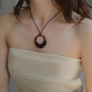 Op de afbeelding: Een ketting met een donkerbruine, cirkelvormige hanger. De glanzende, halvemaanvormige hanger hangt aan een zwart koord. De ketting wordt gedragen over een top van geribbelde stof in neutrale tinten.