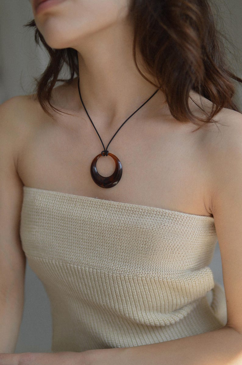 Op de afbeelding: Een ketting met een donkerbruine, cirkelvormige hanger. De glanzende, halvemaanvormige hanger hangt aan een zwart koord. De ketting wordt gedragen over een top van geribbelde stof in neutrale tinten.