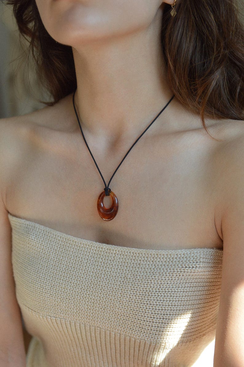 May include: A brown oval pendant necklace with a black cord. The pendant has a layered design and a glossy finish. The necklace is worn around the neck.
