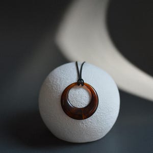 Op de afbeelding: Een bruine hars hanger ketting met een cirkelvormig ontwerp. De hanger hangt aan een zwart koord en rust op een witte, getextureerde bol. De achtergrond is donkergrijs.