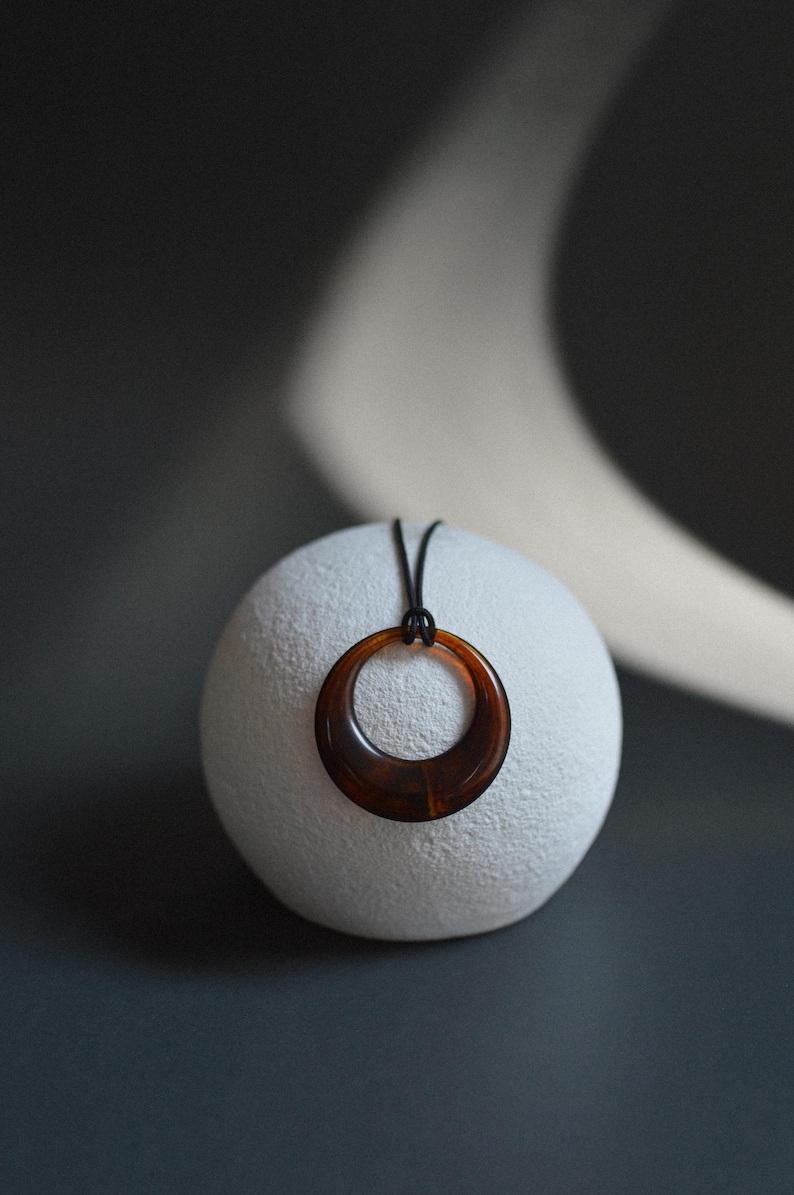 Op de afbeelding: Een bruine hars hanger ketting met een cirkelvormig ontwerp. De hanger hangt aan een zwart koord en rust op een witte, getextureerde bol. De achtergrond is donkergrijs.
