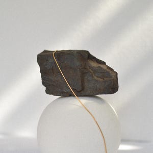 May include: A gold chain necklace draped over a dark grey rock, which is balanced on top of a white sphere.
