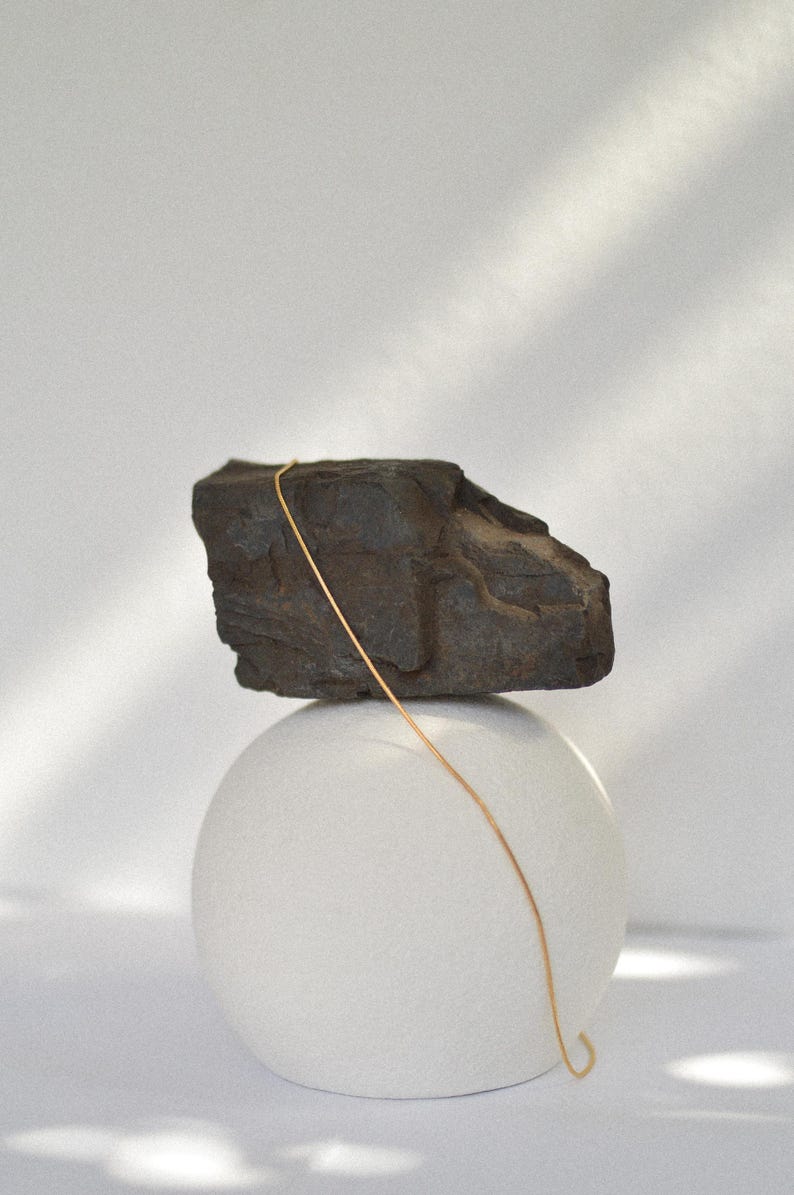 May include: A gold chain necklace draped over a dark grey rock, which is balanced on top of a white sphere.