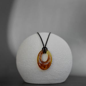 May include: A pendant necklace featuring a double oval pendant in shades of amber and brown, suspended from a black cord. The necklace is displayed on a white, textured, spherical object against a neutral background. Jewelry.