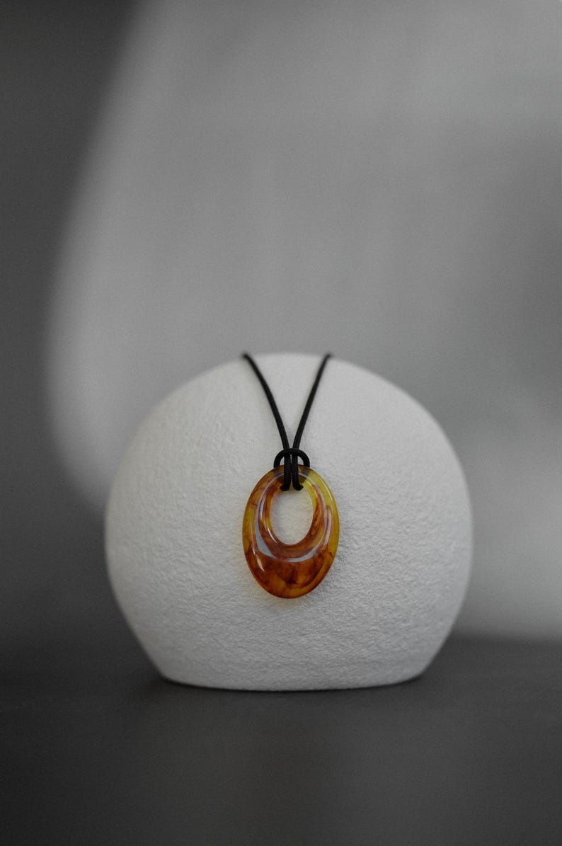 May include: A pendant necklace featuring a double oval pendant in shades of amber and brown, suspended from a black cord. The necklace is displayed on a white, textured, spherical object against a neutral background. Jewelry.